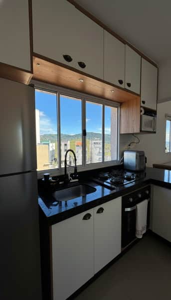 Cozinha compacta com armários brancos e bancada escura. Janela ampla com vista para a cidade.