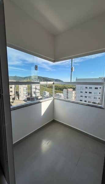 Varanda envidraçada com vista para prédios e morro arborizado. Piso cinza e paredes brancas.