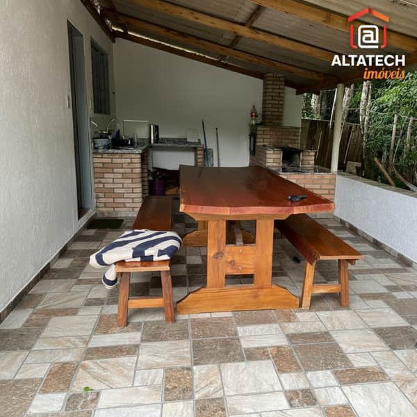 Área externa com churrasqueira, mesa rústica de madeira e bancos. Piso cerâmico e cozinha ao fundo.