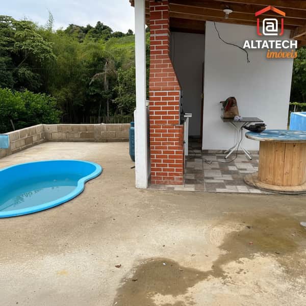 Área externa com piscina, churrasqueira de tijolos e mesa de madeira. Paisagem verde ao fundo.