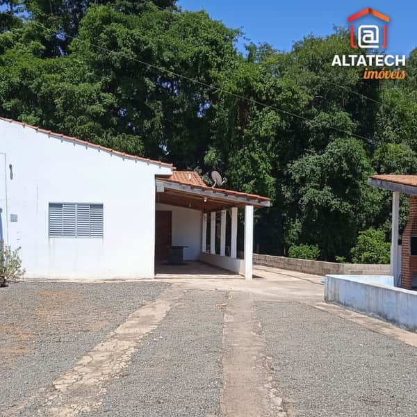 Fachada de casa térrea branca com varanda e área externa com piso de pedra e vegetação ao fundo.