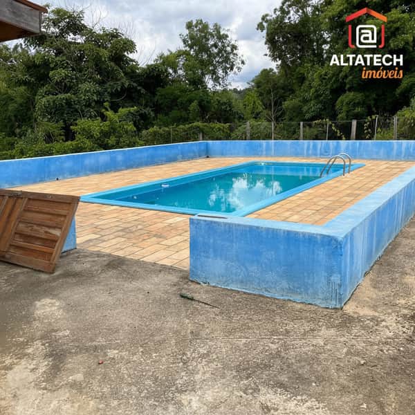 Área externa com piscina retangular e piso de tijolos. Muro azul ao redor. Vegetação ao fundo.
