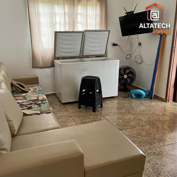 Sala com sofá bege, freezer horizontal aberto, TV e piso cerâmico. Ambiente amplo e iluminado.
