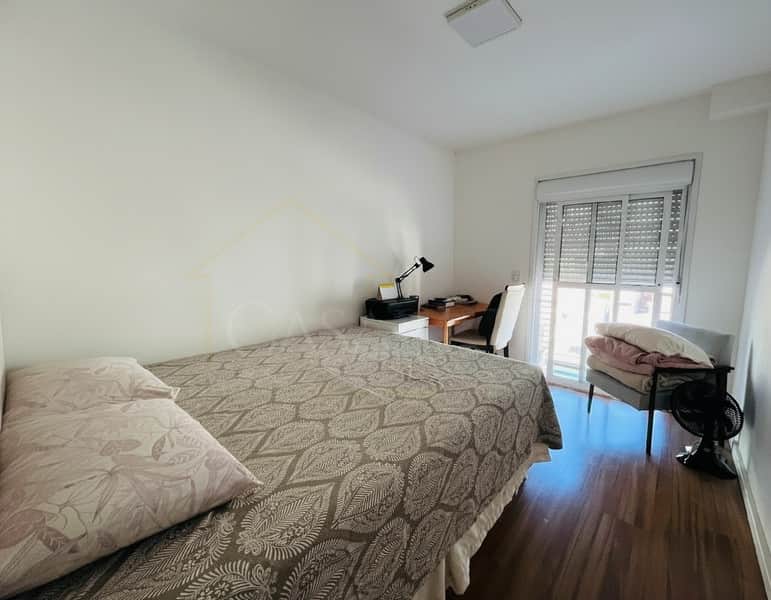 Quarto com cama de casal, escrivaninha, cadeira e poltrona perto da janela com persiana. Piso de madeira.