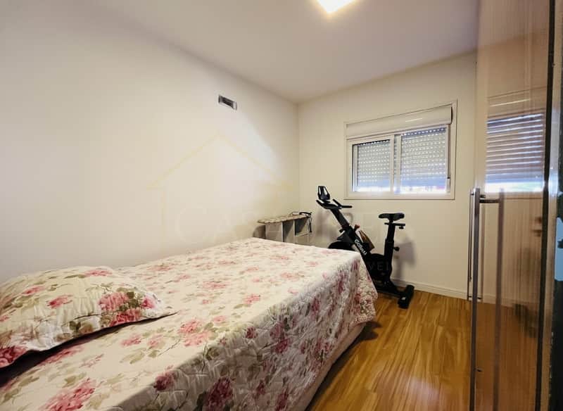 Quarto com cama florida, bicicleta ergométrica, janela com persiana e piso de madeira clara.