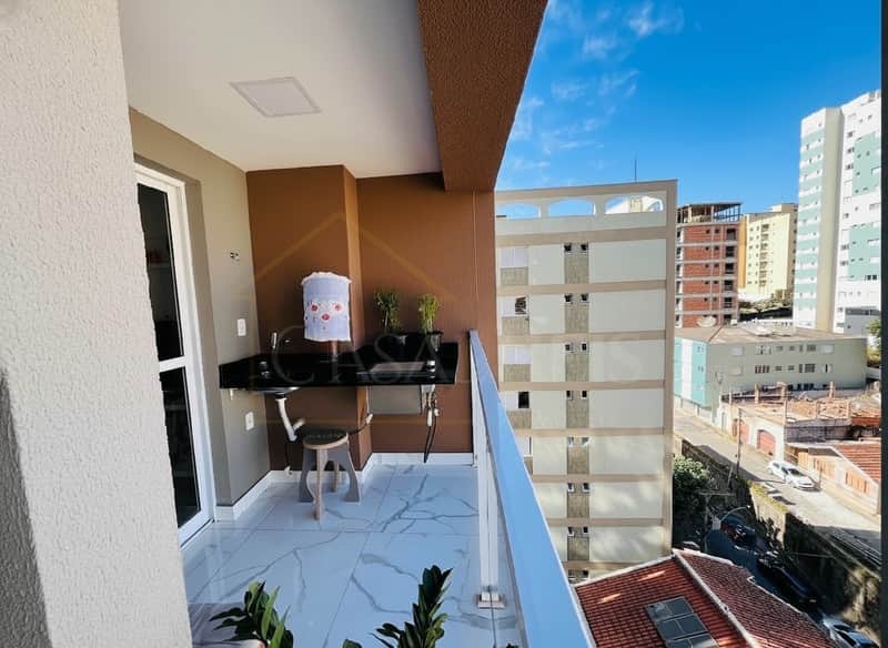 Varanda gourmet com pia, bancada preta e piso claro. Vista para prédios e rua arborizada.