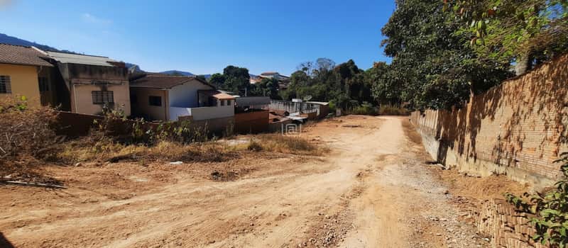 Terreno baldio em área urbana. Muro de tijolos à direita, casas ao fundo. Céu azul.