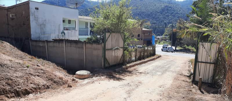 Área externa com portão, muro e vegetação. Rua com carro ao fundo e montanhas na paisagem.