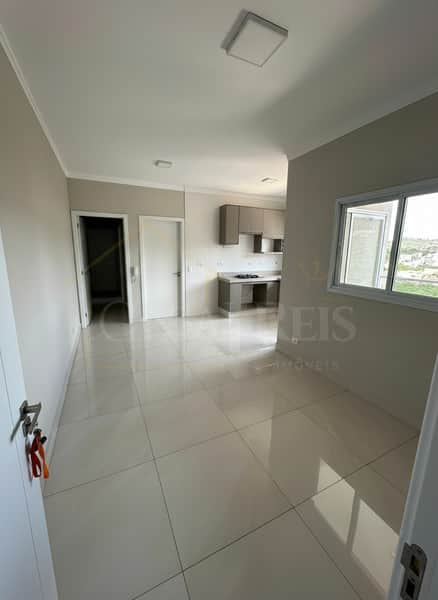 Sala clara com piso cerâmico, janela e cozinha americana ao fundo. Apartamento novo.