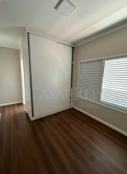 Quarto com piso amadeirado, armário embutido branco e janela com persianas claras. Ambiente clean e iluminado.
