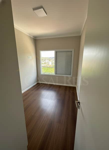 Quarto claro com piso amadeirado e janela com vista para a cidade. Ambiente arejado e iluminado.