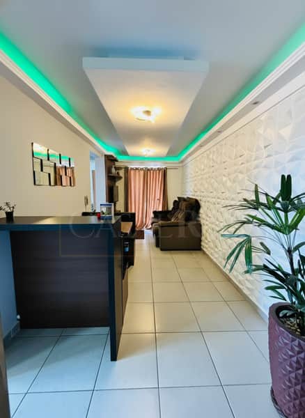 Sala decorada com painel 3D, iluminação verde no teto e móveis escuros. Piso claro e planta decorativa.