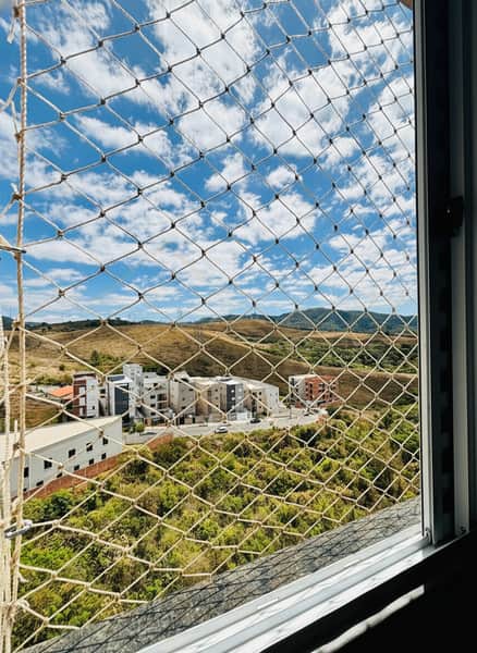 Vista da janela com rede de proteção. Prédios, morros e céu azul com nuvens ao fundo.
