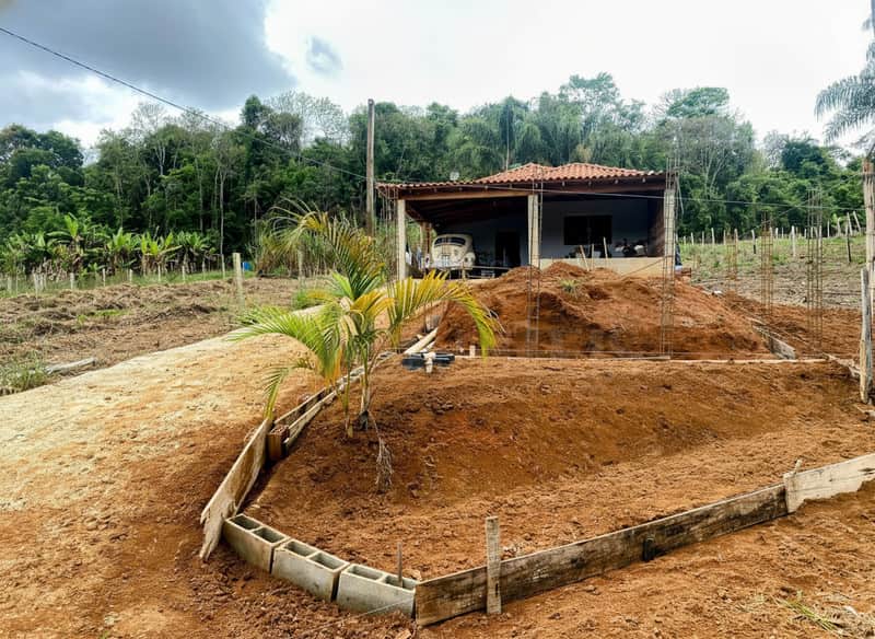 Área externa com casa ao fundo. Terreno em construção, terra batida e vegetação.