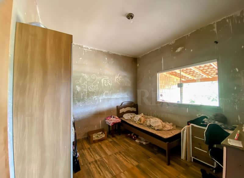Quarto com cama de solteiro, guarda-roupa e paredes inacabadas. Piso amadeirado e janela com vista externa.