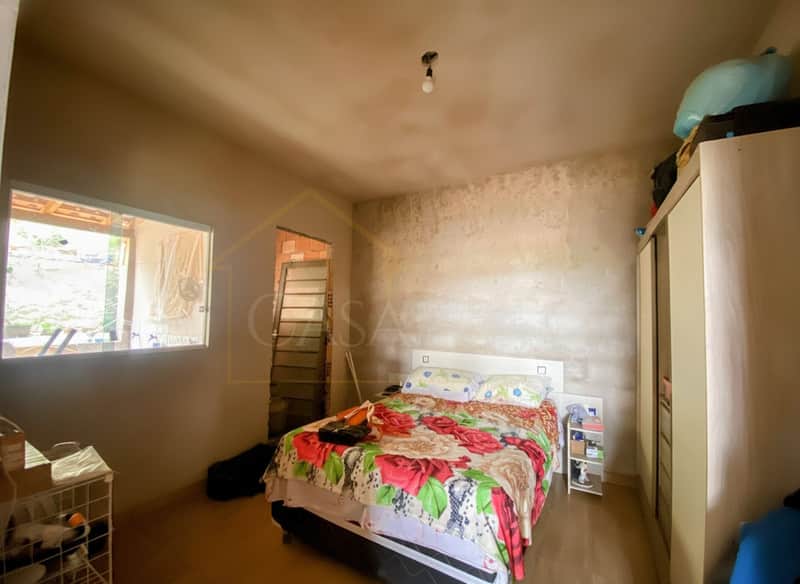Quarto com cama floral, guarda-roupa claro e janela. Paredes em tom bege e piso amadeirado.