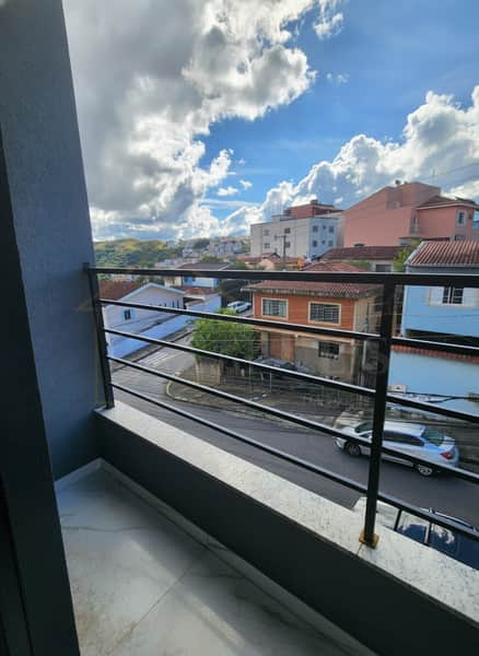 Varanda com vista urbana. Guarda-corpo preto, piso claro. Ao fundo, casas coloridas e céu azul com nuvens.