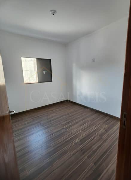 Quarto vazio com piso amadeirado e janela. Paredes brancas e iluminação central. Espaço arejado e pronto para mobiliar.