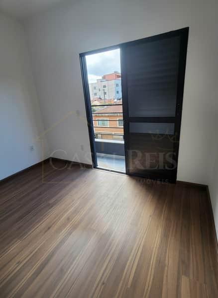 Quarto com piso amadeirado e porta para varanda com vista urbana. Ambiente claro e arejado.