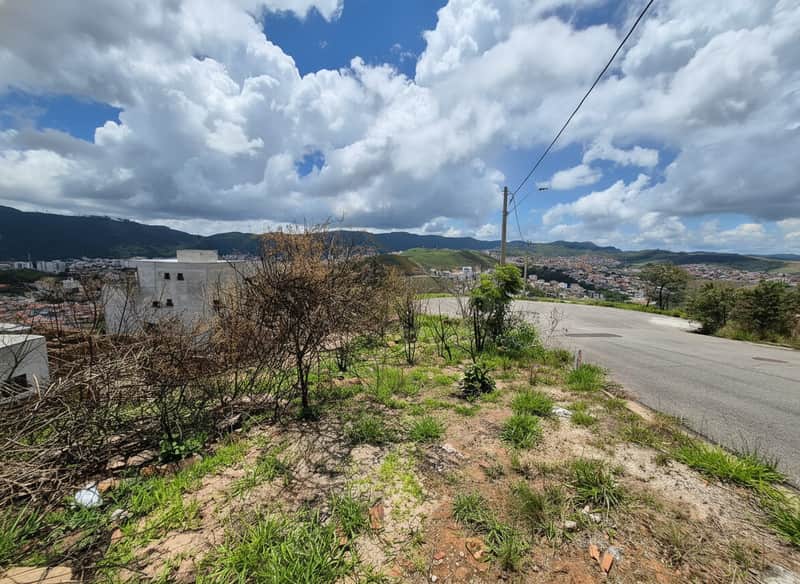 Terreno em declive com vegetação rasteira e vista para a cidade e montanhas. Céu com nuvens.