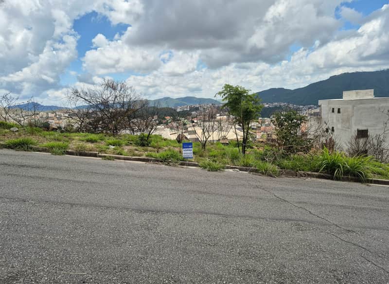 Terreno à venda com vista panorâmica da cidade e montanhas, vegetação rasteira e placa de imobiliária.