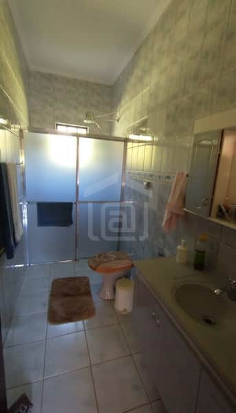 Banheiro com azulejos claros, box de vidro fosco, pia com gabinete e vaso sanitário com tampa laranja.