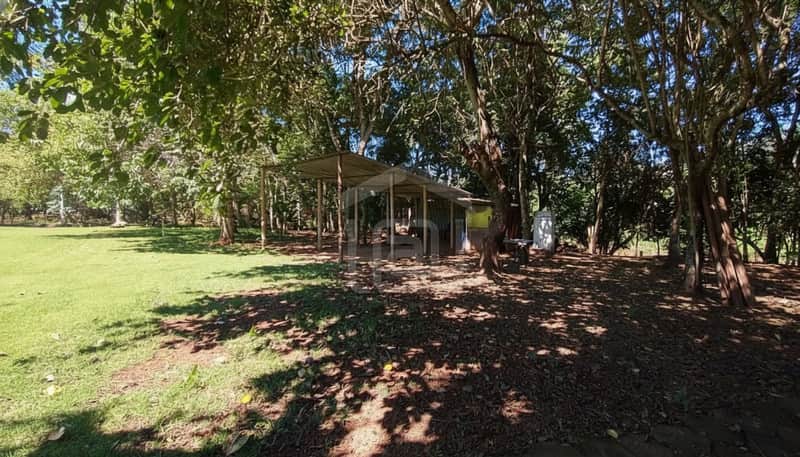 Área externa ensolarada com gramado, árvores e estrutura metálica coberta. Ambiente campestre e tranquilo.