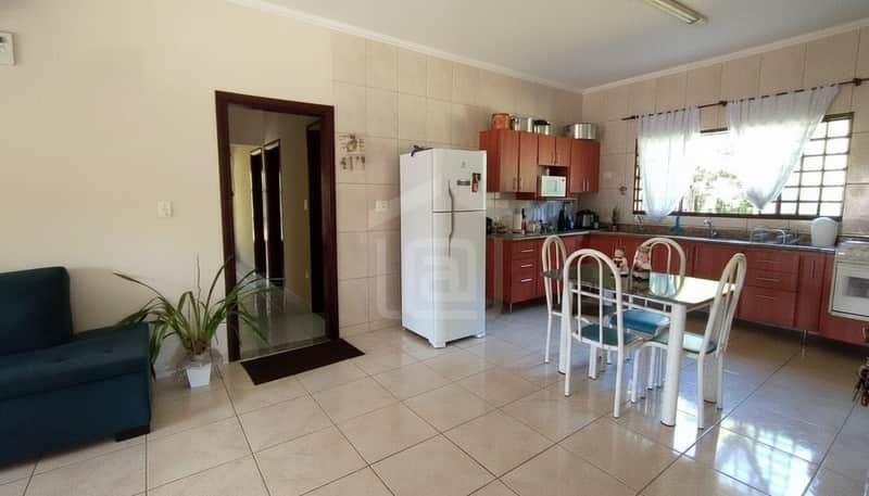 Cozinha ampla com armários em madeira, mesa com cadeiras e geladeira branca. Ambiente claro e arejado.