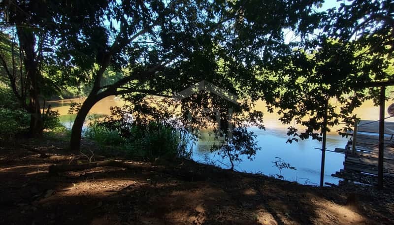 Área externa arborizada com rio calmo e deck de madeira. Natureza exuberante e sombra fresca.