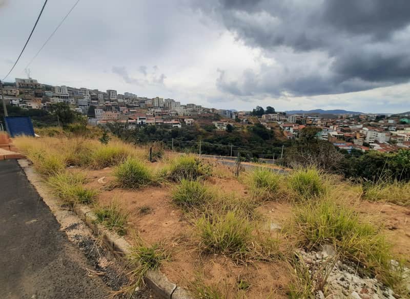 Terreno com vegetação rasteira e vista para casas na encosta sob céu nublado. Área externa com potencial construtivo.