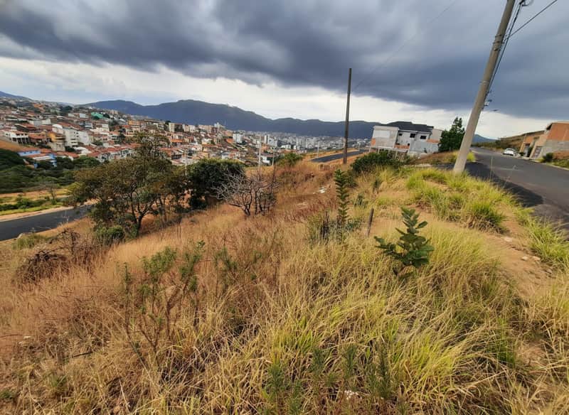 Terreno em declive com vegetação rasteira e vista para a cidade. Céu nublado e rua asfaltada ao lado.