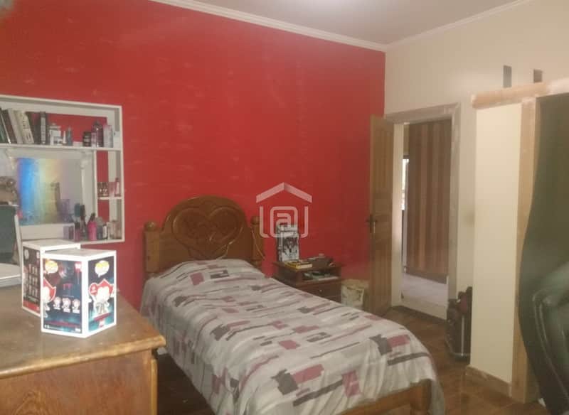 Quarto com cama de madeira, parede vermelha, estante e cômoda. Decoração simples e aconchegante.
