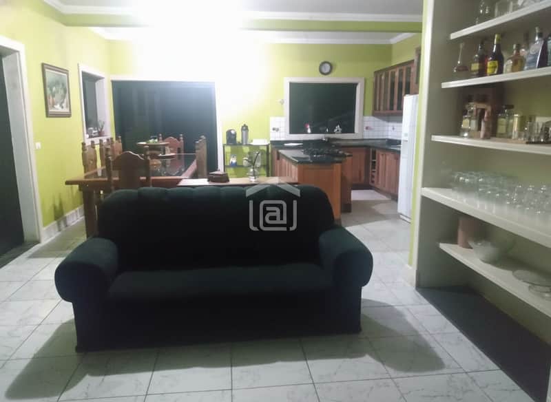 Sala ampla com sofá preto, mesa de jantar e cozinha americana com armários de madeira. Iluminação suave.