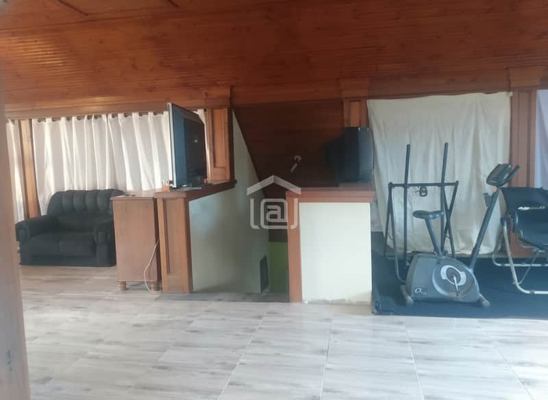 Sala ampla com teto de madeira, TV, sofá escuro e equipamentos de ginástica. Piso claro.