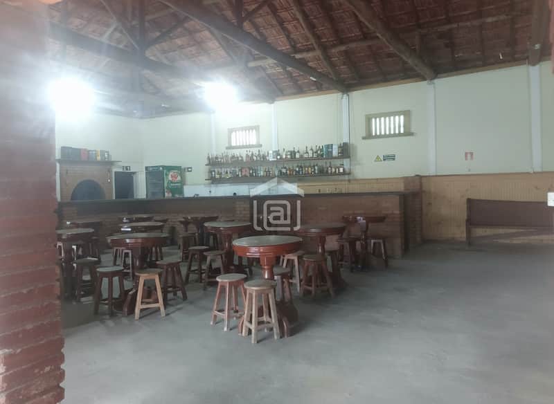 Salão amplo com mesas e cadeiras de madeira, bar com prateleiras de bebidas e teto rústico.