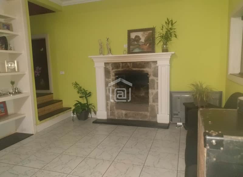 Sala verde com lareira rústica, estante branca e piso cerâmico claro. Ambiente iluminado e aconchegante.