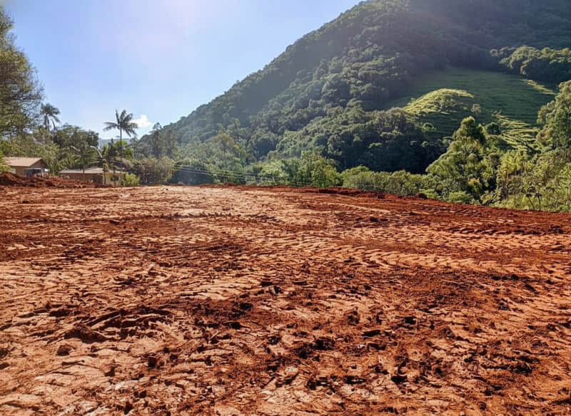 Terreno plano de terra batida com marcas de pneus e montanha verde ao fundo. Área externa para construção.