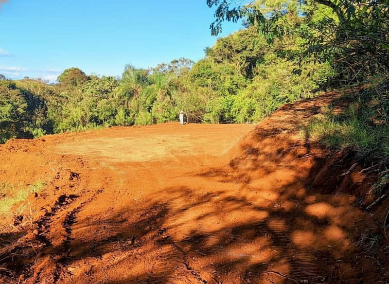 Área externa ampla com terra vermelha e vegetação ao fundo. Espaço preparado para construção.