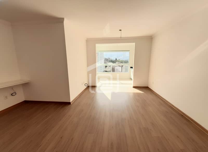 Sala clara com piso amadeirado e janela ampla, vista para área externa com prédios. Ambiente arejado.