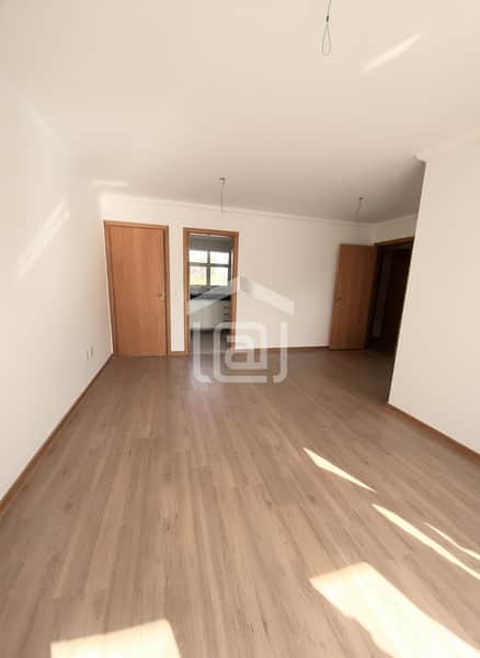 Sala ampla com piso amadeirado, paredes brancas e portas de madeira clara. Ambiente iluminado e arejado.