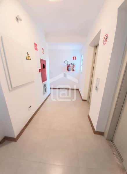Corredor claro com piso cerâmico, extintores e sinalização de segurança. Elevador à direita.