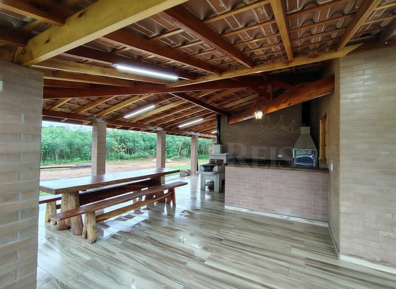 Área externa com churrasqueira, mesa rústica e piso cerâmico. Cobertura de madeira e tijolos aparentes.
