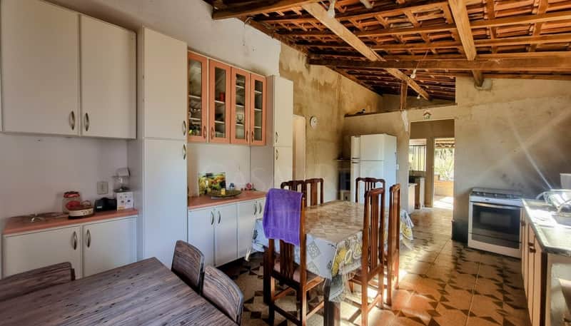 Cozinha rústica com armários brancos e mesa de jantar em madeira. Piso cerâmico e teto aparente com vigas.