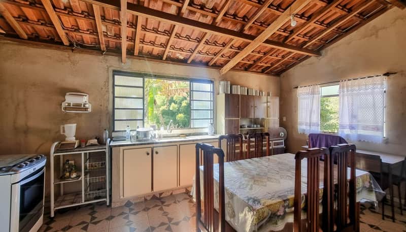 Cozinha rústica com mesa, armários antigos e janela ampla. Piso decorado e teto de madeira aparente.
