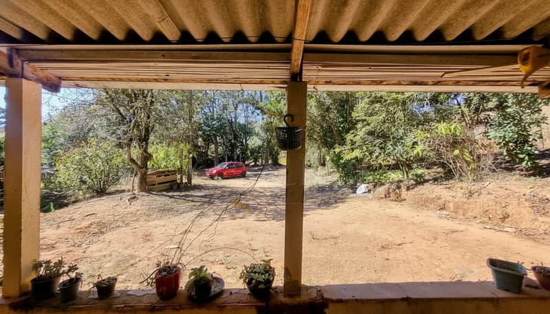 Varanda rústica com vista para área externa arborizada e carro vermelho ao fundo. Plantas decoram a varanda.