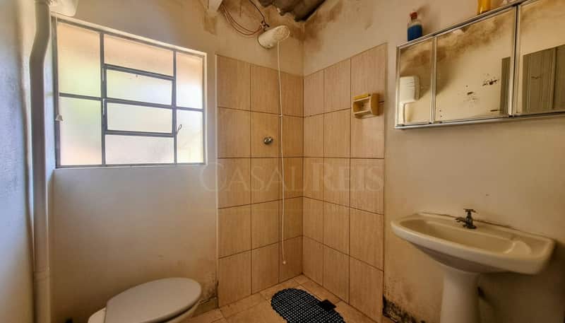 Banheiro antigo com pia, vaso sanitário, box com azulejos bege e armário com espelho sujo. Janela com luz natural.