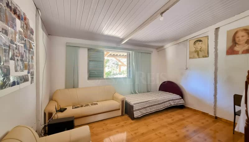 Quarto aconchegante com cama, sofá bege, piso cerâmico e iluminação natural da janela. Decoração simples.