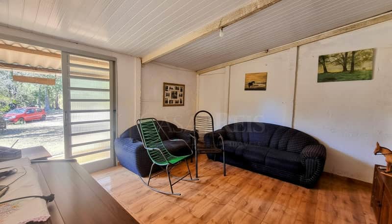 Sala rústica com sofás pretos, cadeiras de vime e piso de madeira. Porta de vidro com vista para área externa.