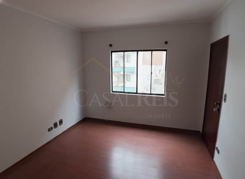 Quarto vazio com piso de madeira escura, janela e porta marrom. Paredes brancas e tomadas.