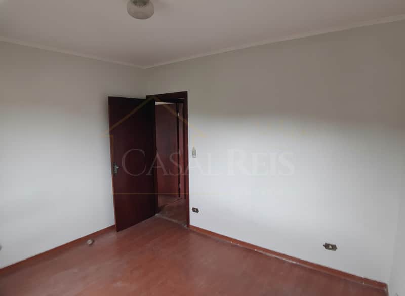 Quarto vazio com piso de madeira escura e porta aberta. Paredes brancas e iluminação central.